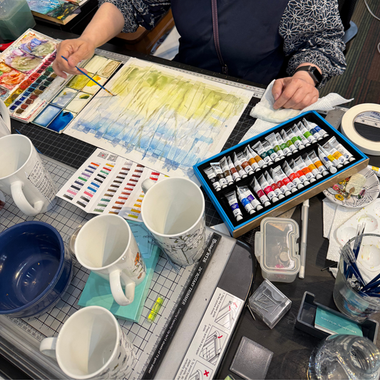 Artistic workspace with watercolor paints, brushes, and mugs on a table.